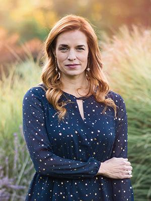 White woman with red hair standing in a field wearing a deep teal blouse with gold stars